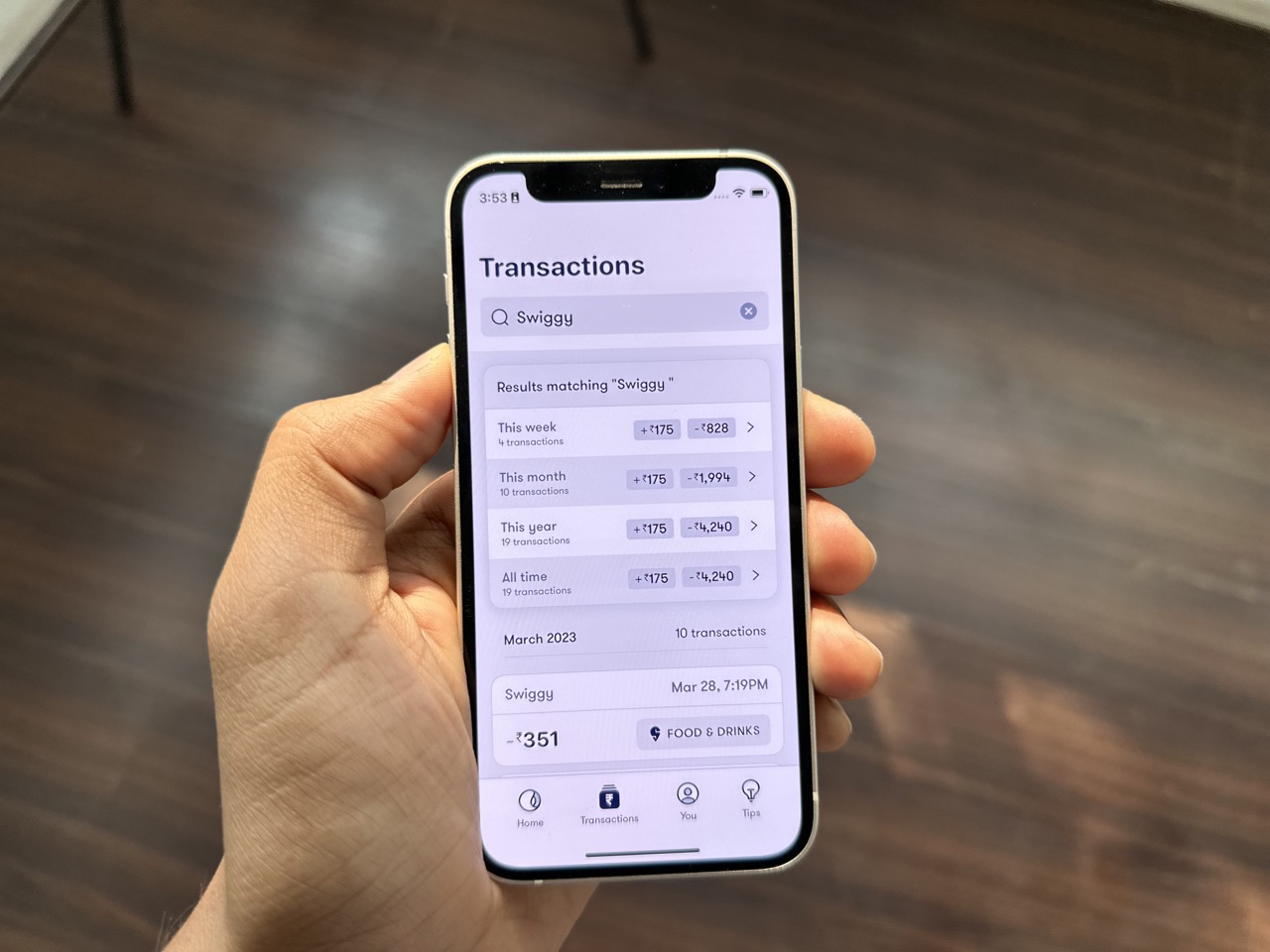 A person holding the iphone with Fold app open. In the app - "swiggy" is in transaction search field and in results there is a summary which shows transactions in this week, this month, this year, and all time with amounts.